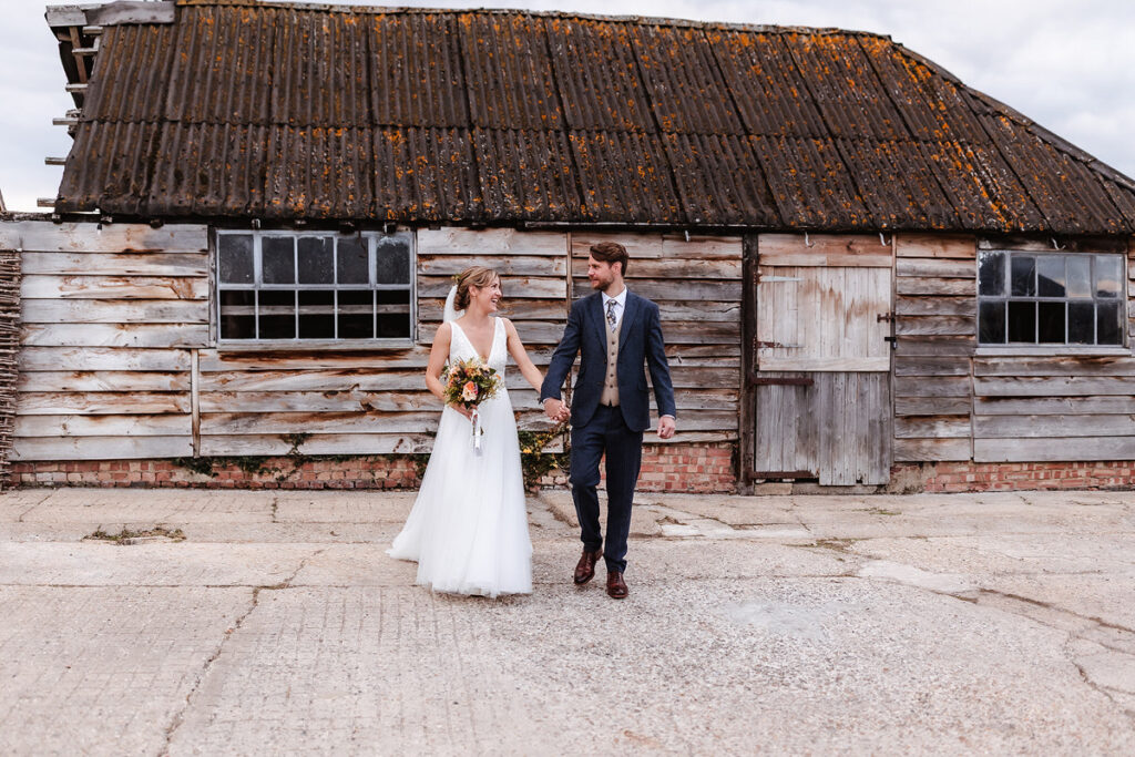 A bride in a white dress holding a bouquet and a groom in a suit hold hands while walking in front of an old, rustic wooden barn with weathered boards and a corrugated roof.