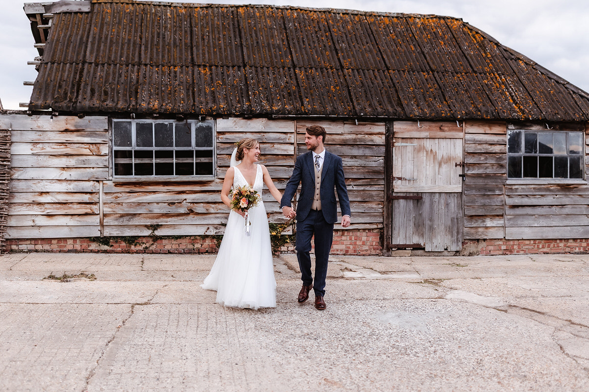 A bride in a white dress holding a bouquet and a groom in a suit hold hands while walking in front of an old, rustic wooden barn with weathered boards and a corrugated roof.