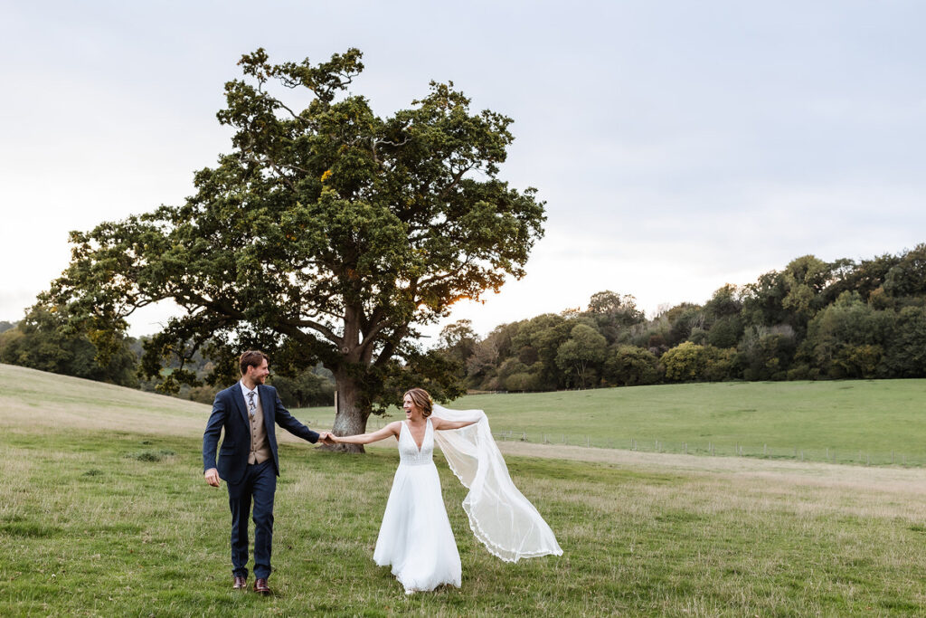 A bride and groom walk holding hands in a grassy field, smiling at each other. The bride’s veil flows in the breeze. A large tree and rolling hills are in the background under a cloudy sky.