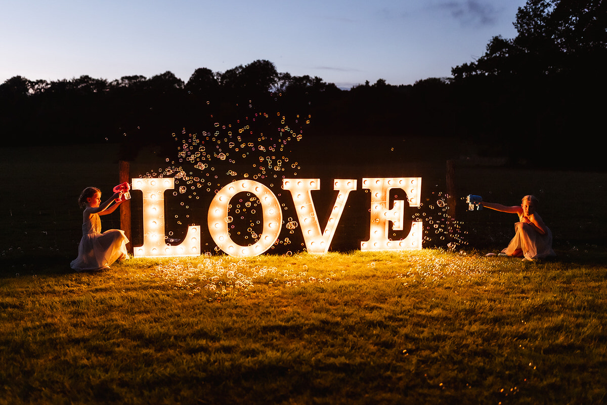 Two children blow bubbles on either side of large, illuminated letters spelling LOVE outdoors at dusk, with trees silhouetted in the background.