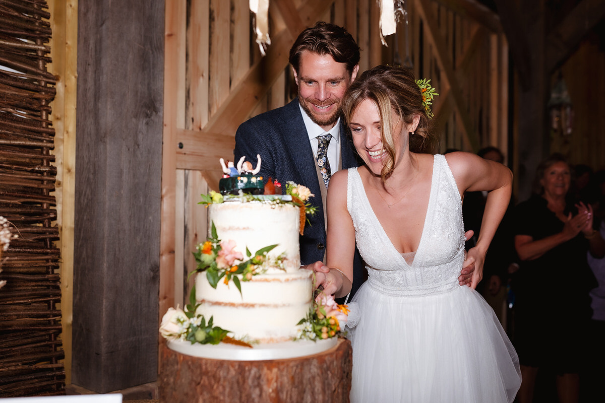 A bride and groom smile and cut a rustic, decorated wedding cake together at their reception in a wooden barn-like setting. The bride wears a white dress; the groom is in a suit.