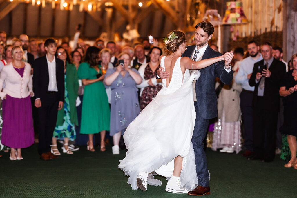 A bride in a white dress and sneakers dances with the groom in a suit at a wedding reception, surrounded by guests watching and taking photos in a warmly lit venue.