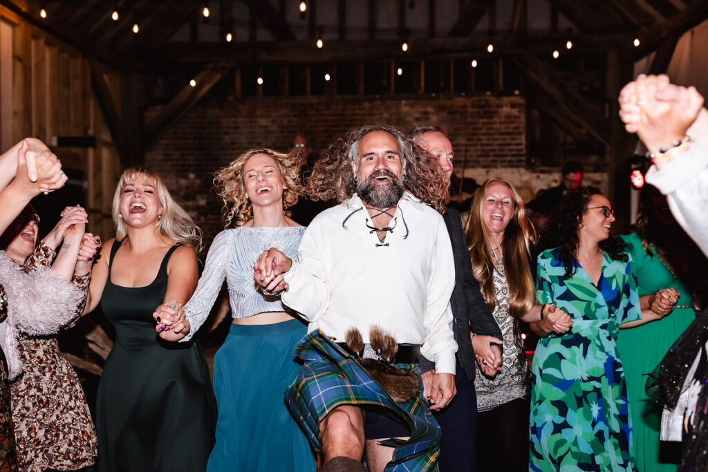 A group of people dance energetically in a barn-style venue, holding hands and smiling. The man in front wears a kilt and white shirt, leading the cheerful group under string lights.