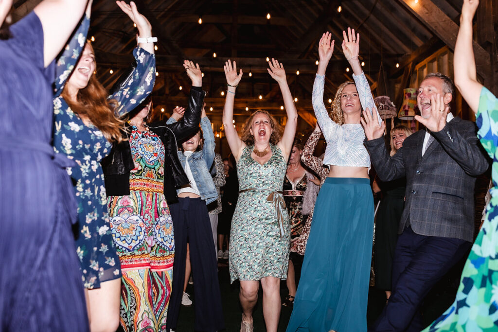 A group of people dressed in colorful outfits are dancing enthusiastically with raised arms at a lively indoor event, possibly a wedding or party, under string lights in a rustic wooden venue.
