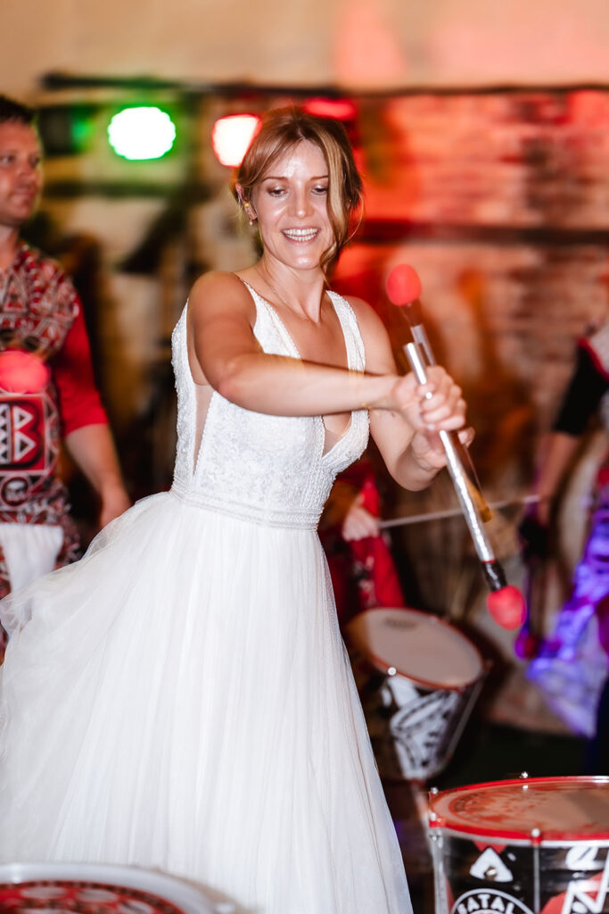 A woman in a white wedding dress smiles while playing a drum with red mallets at a lively event, with colorful lights and other drummers in the background.