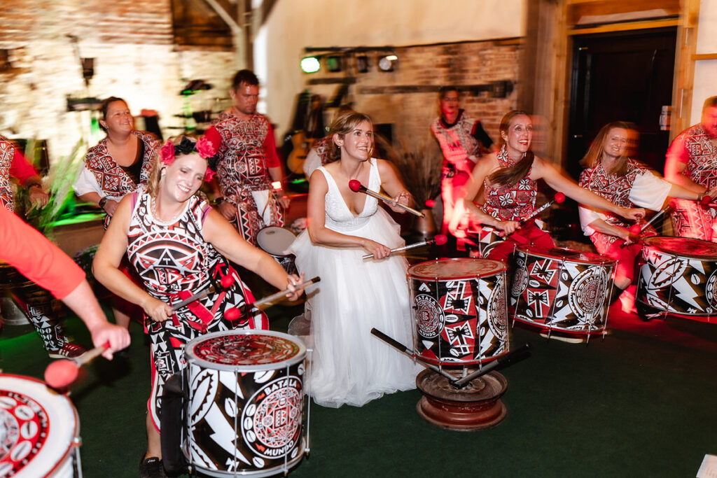 A bride in a white dress joyfully plays drums alongside a lively group of drummers dressed in matching red, white, and black outfits at an indoor event. Everyone is smiling and engaged in the music.