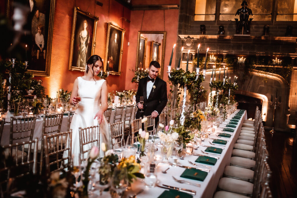 A bride and groom stand by a long, elegant dining table set for a formal event in a grand hall with portraits, candles, and floral arrangements—captured beautifully by a Hampshire wedding photographer.