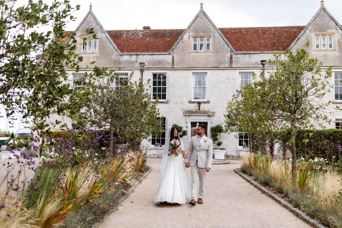 A bride and groom in light-colored wedding attire walk hand in hand along a garden path before a grand, historic manor house. Trees and flowers line the way, beautifully captured by a Hampshire wedding photographer.