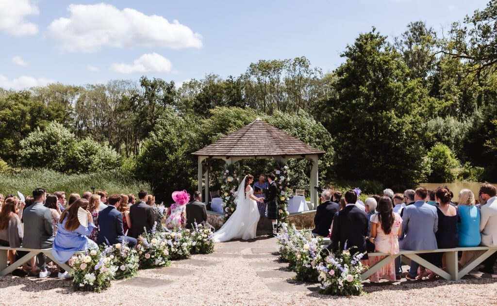 A bride and groom stand under a wooden gazebo during an outdoor wedding ceremony. Guests sit on benches lined with flower arrangements, surrounded by greenery and trees on a sunny day.
