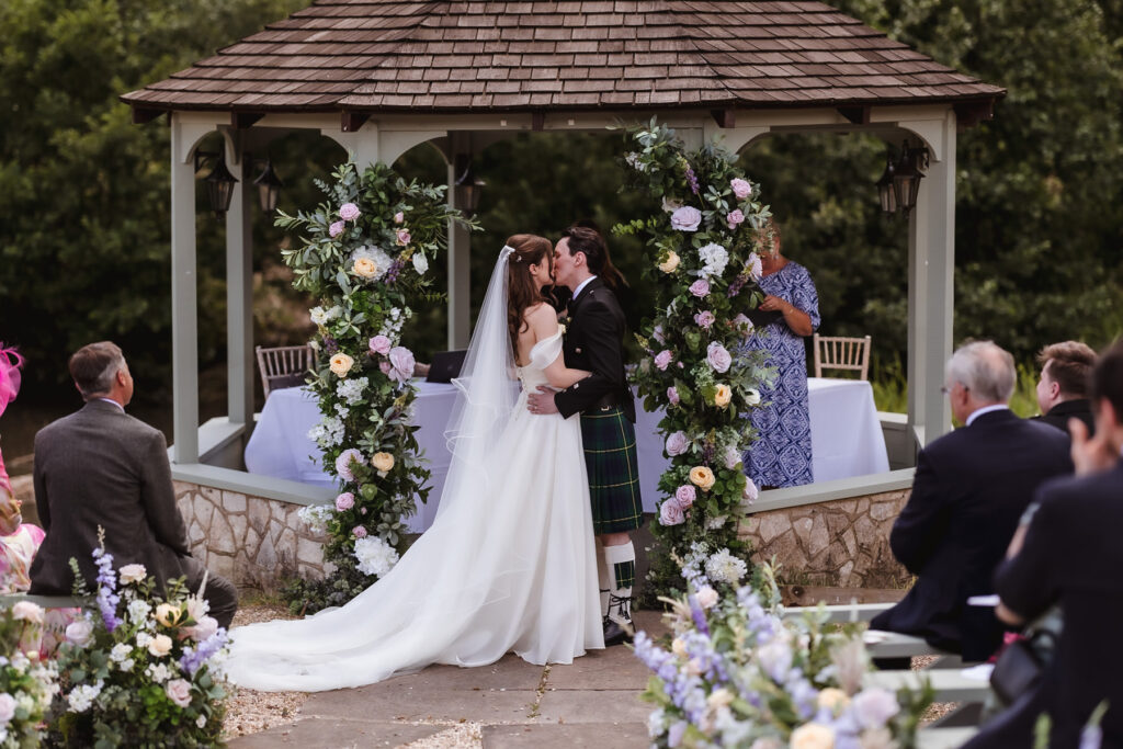 A bride and groom share a kiss under a flower-adorned gazebo during an outdoor wedding ceremony, surrounded by guests seated on benches. The groom wears a kilt and the bride is in a white gown with a long train.