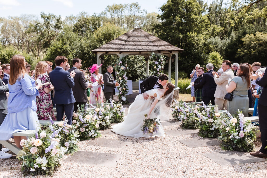 A bride and groom kiss as the groom dips the bride in the center aisle of an outdoor wedding, surrounded by guests clapping and flower arrangements, with a gazebo and trees in the background.
