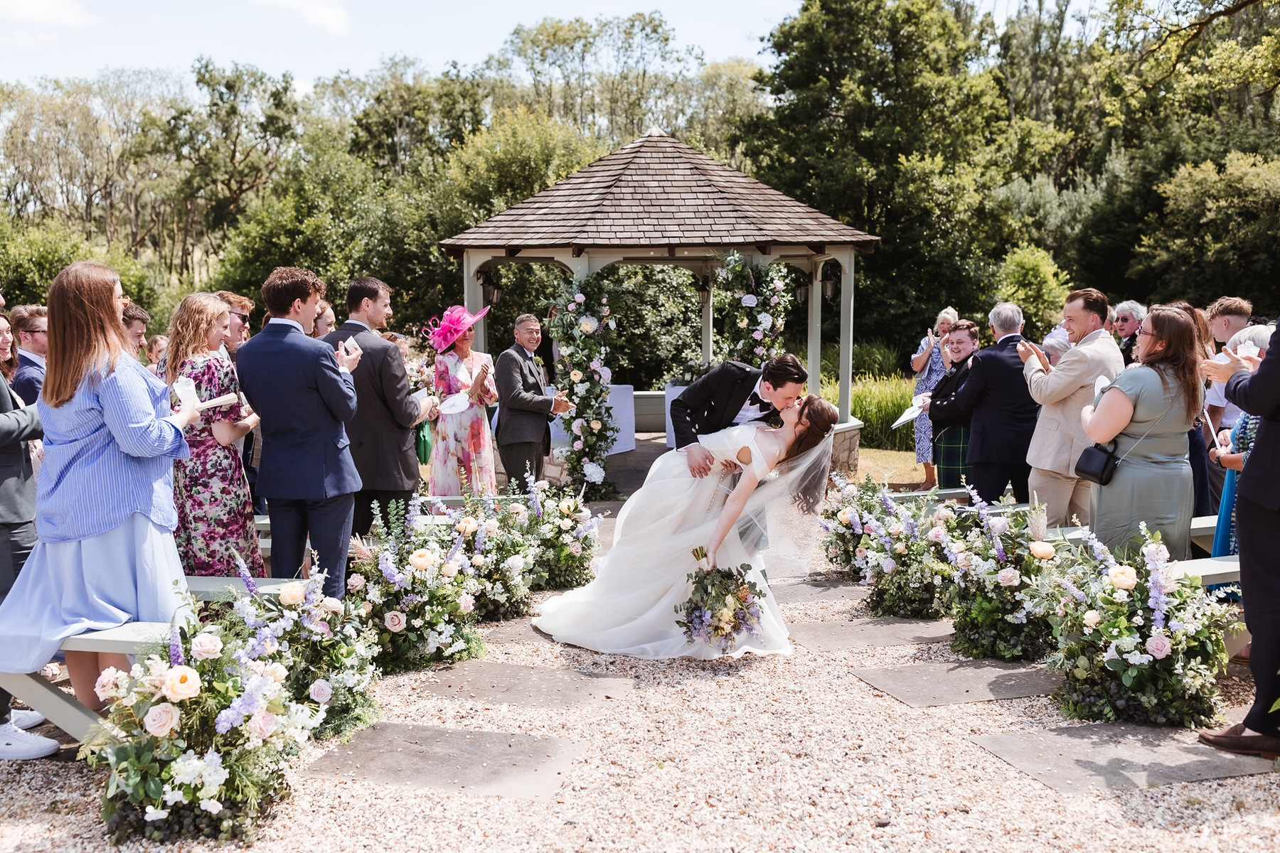 A bride and groom kiss as the groom dips the bride in the center aisle of an outdoor wedding, surrounded by guests clapping and flower arrangements, with a gazebo and trees in the background.