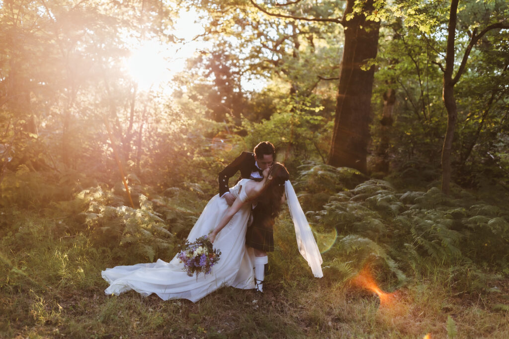 A groom in traditional Scottish attire dips his bride in a forest clearing at sunset. Captured by a Hampshire wedding photographer, the bride’s veil flows behind her as sunlight filters through the trees, creating a warm, romantic atmosphere.