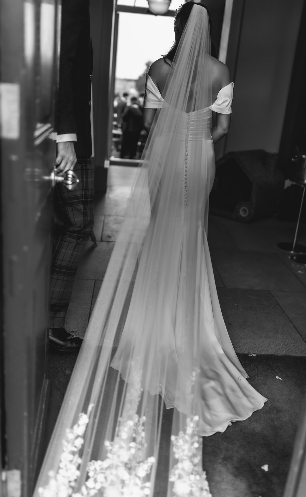 A bride in an off-the-shoulder gown with a long veil walks indoors beside someone in a suit. Captured by a Hampshire wedding photographer, this black and white photo is taken from behind as light streams in from an open door ahead.