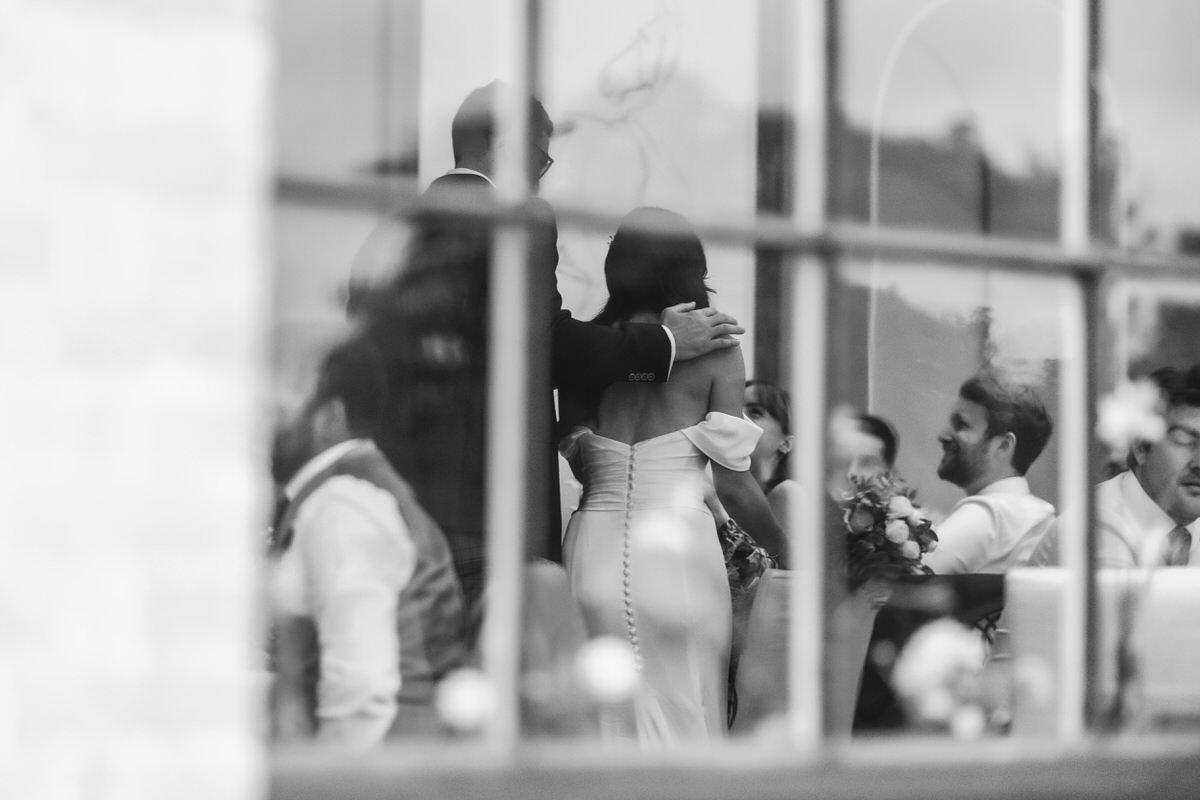 A black and white photo taken through a window by a Hampshire wedding photographer shows a couple embracing among guests at a formal event. The scene appears joyful, with people smiling and holding flowers.
