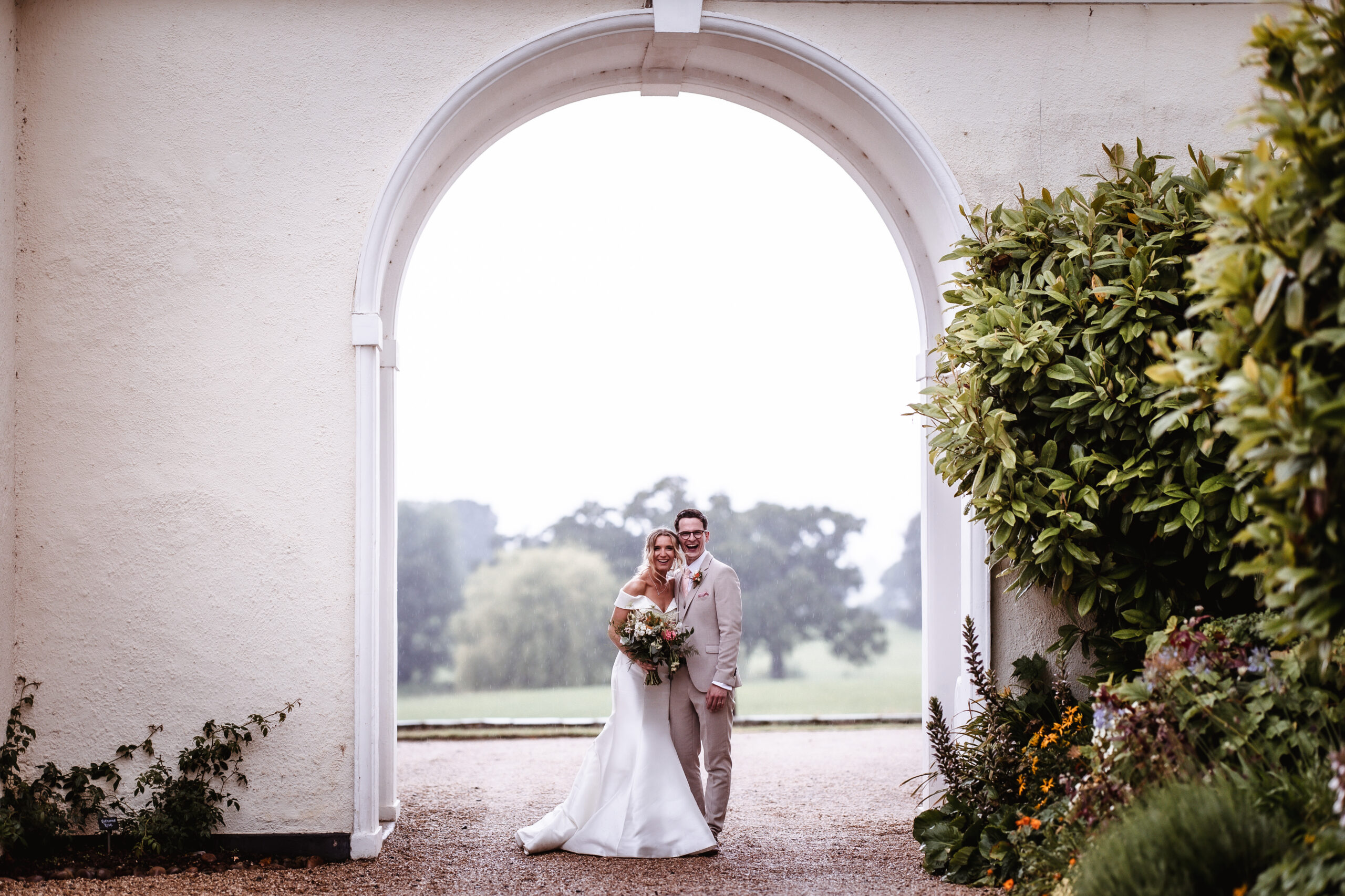A bride and groom stand smiling under a large white archway, surrounded by greenery and flowers, with a scenic landscape in the background.
