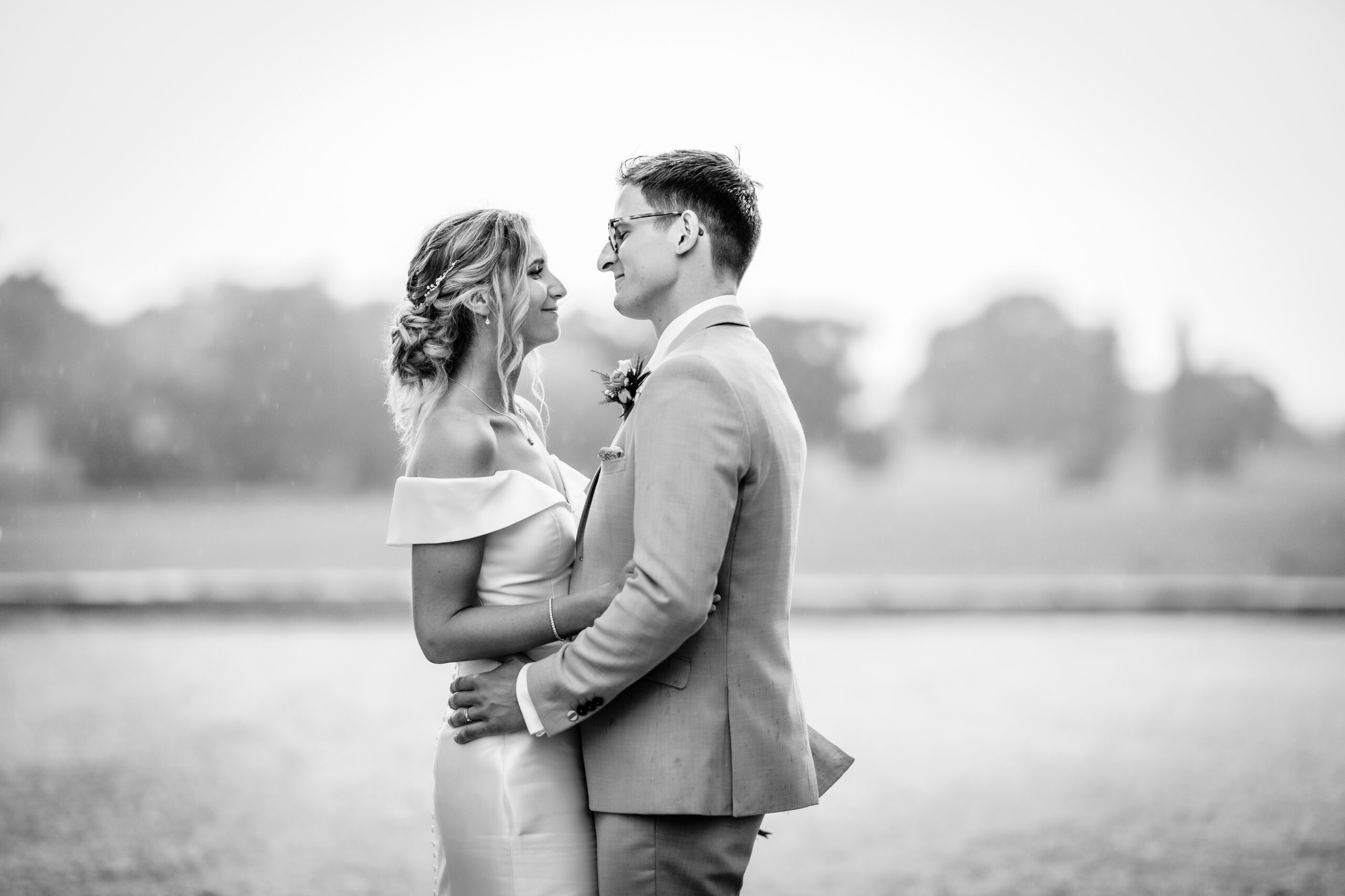 A bride and groom stand closely together outdoors, gazing into each others eyes and smiling. The background is blurred, highlighting the couple. The image is in black and white.