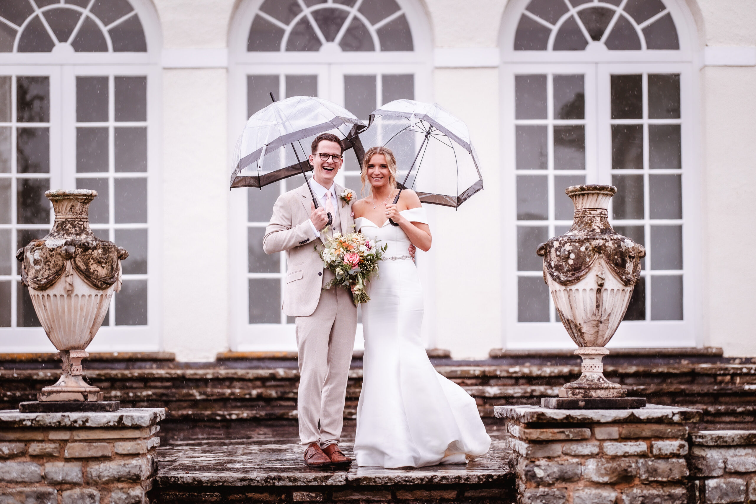A bride and groom stand smiling on stone steps, each holding a clear umbrella in the rain. The bride wears a white dress and holds a bouquet; the groom wears a beige suit. Large urns and arched windows are behind them.