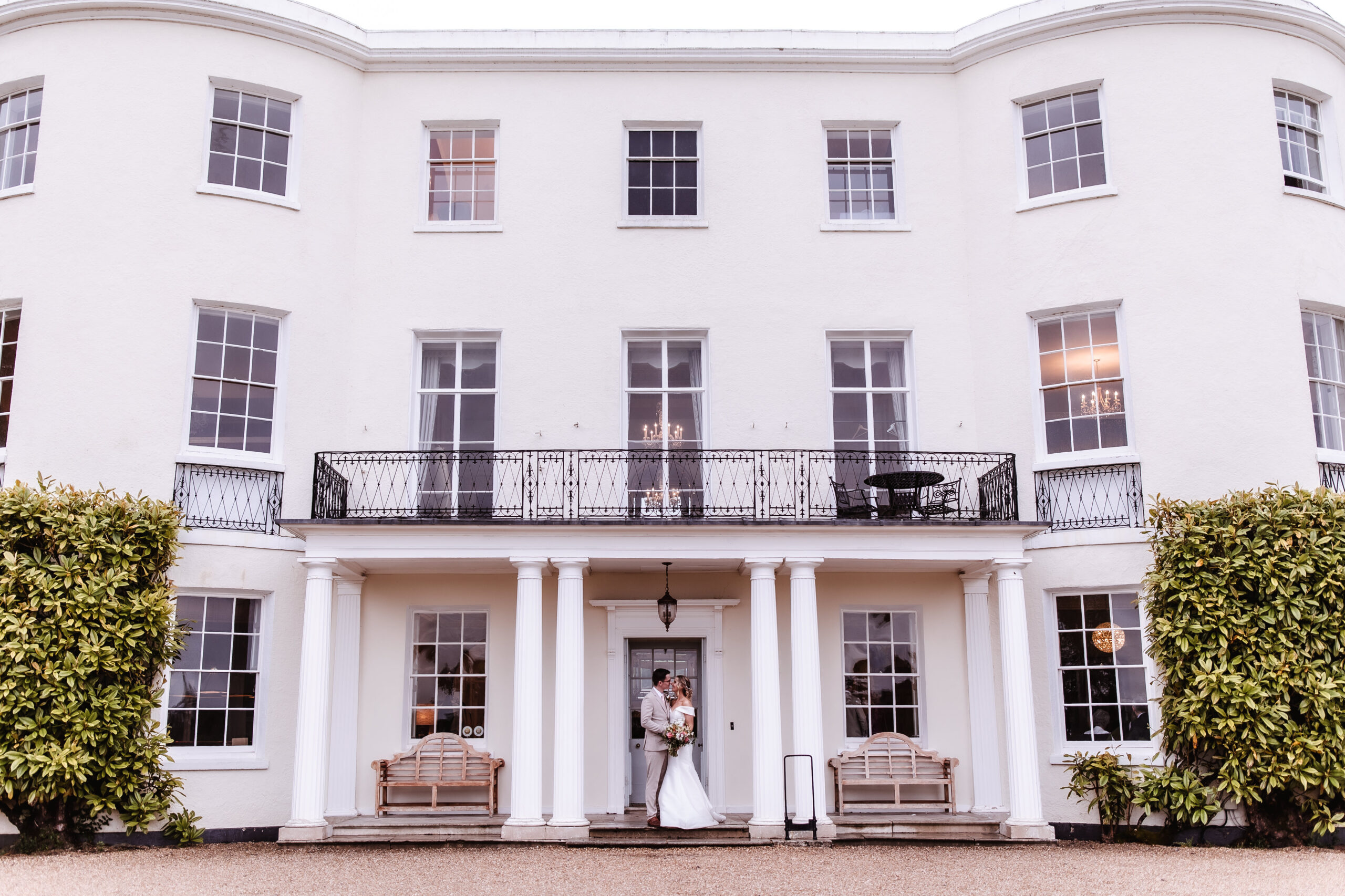 A bride and groom stand together in front of a large, white, two-story building with tall windows, columns, and greenery on either side of the entrance.