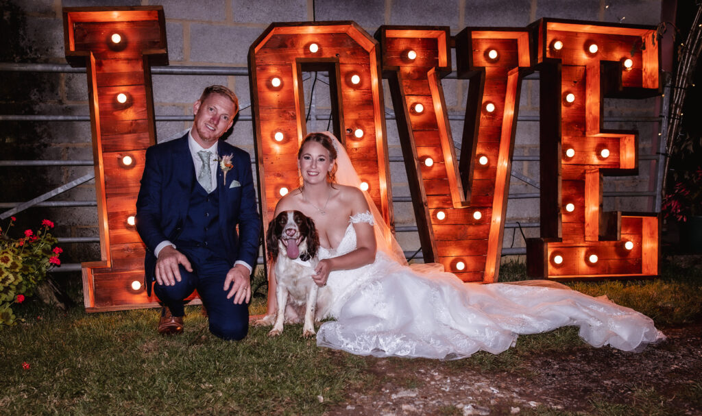 A bride and groom in wedding attire kneel with a dog in front of large, illuminated letters spelling LOVE outdoors at night.