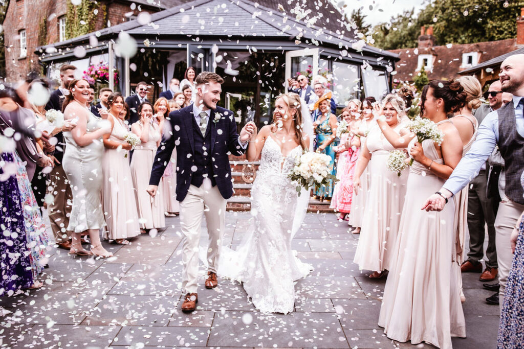 A bride and groom walk hand in hand, smiling, as guests shower them with white confetti outside a venue. The wedding party, dressed in light colors, celebrates around them.
