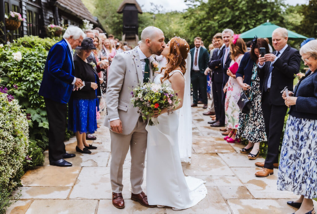 A bride and groom share a kiss outdoors, surrounded by smiling guests in formal attire. The bride holds a bouquet, and petals are scattered on the ground. The scene is joyful and festive.