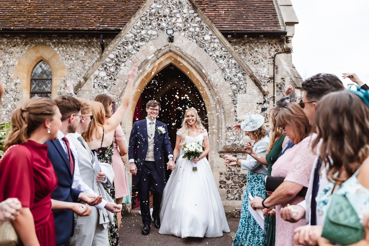 A newlywed couple exits a stone church as guests cheer and throw confetti. The bride, in a white dress holding a bouquet, and the groom, in a suit, smile while surrounded by friends and family celebrating.