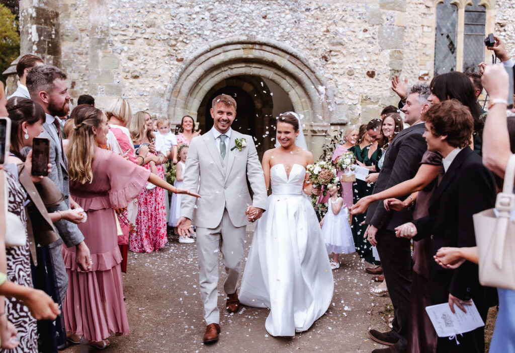 A smiling bride and groom walk hand in hand outside a stone church as guests shower them with confetti. Friends and family cheer and take photos, creating a joyful and celebratory atmosphere.