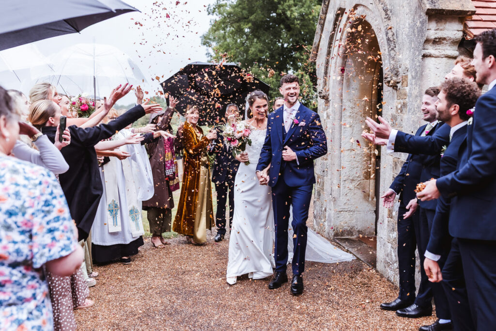 A newlywed couple walks joyfully out of a church as guests shower them with confetti. Both are smiling and holding hands, surrounded by friends and family in formal attire, some holding umbrellas.