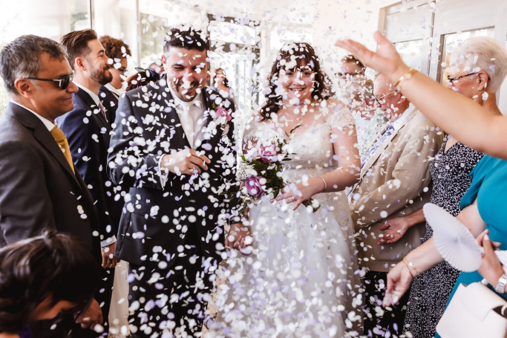 A bride and groom smile as guests shower them with white confetti at their wedding, surrounded by friends and family in celebratory attire.