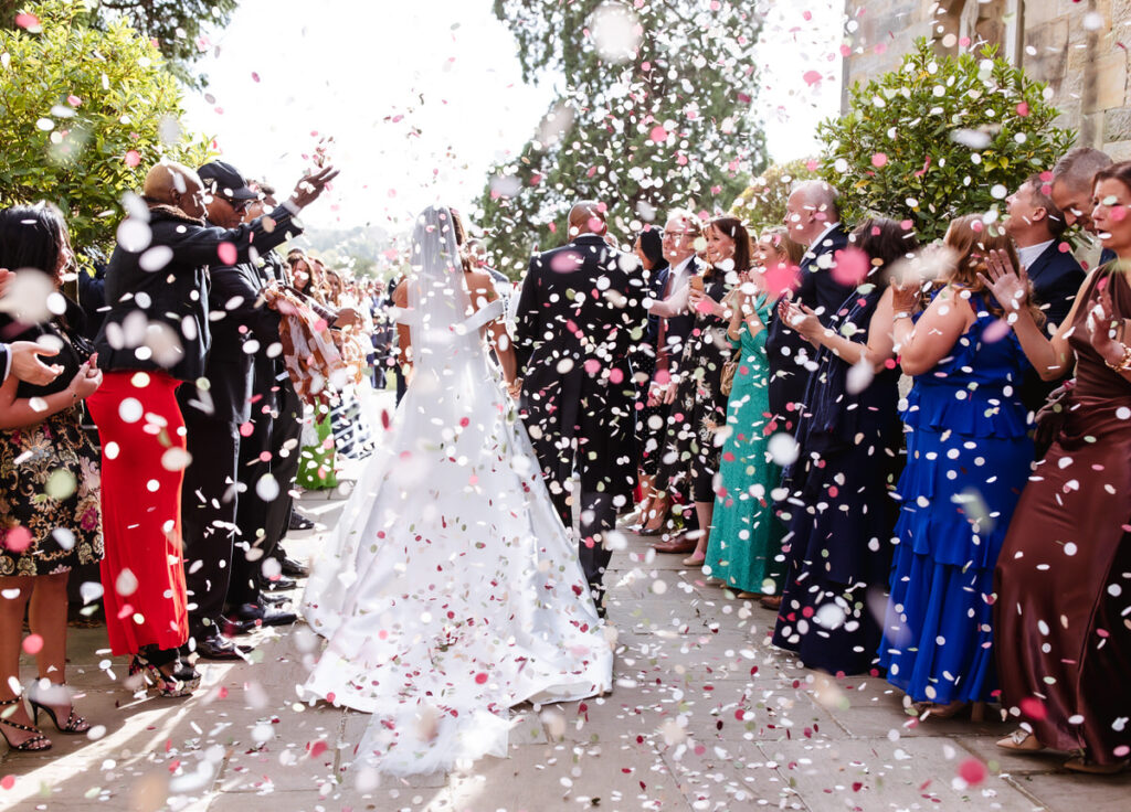A bride and groom walk outside among guests who are throwing pink and white confetti. The couple is framed by smiling, applauding people in colorful dresses and suits on a sunny day.