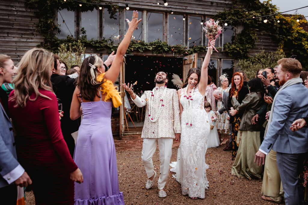 A newly married couple walks joyfully outside as guests shower them with confetti. The bride holds a bouquet and wears a lace dress, while the groom smiles in a cream suit. Friends and family cheer around them in colorful outfits.