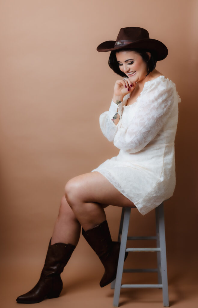 A woman wearing a white dress, dark cowboy hat, and brown boots sits on a stool against a beige background, smiling and looking down with her hand near her chin—a relaxed style for Hampshire business headshots.