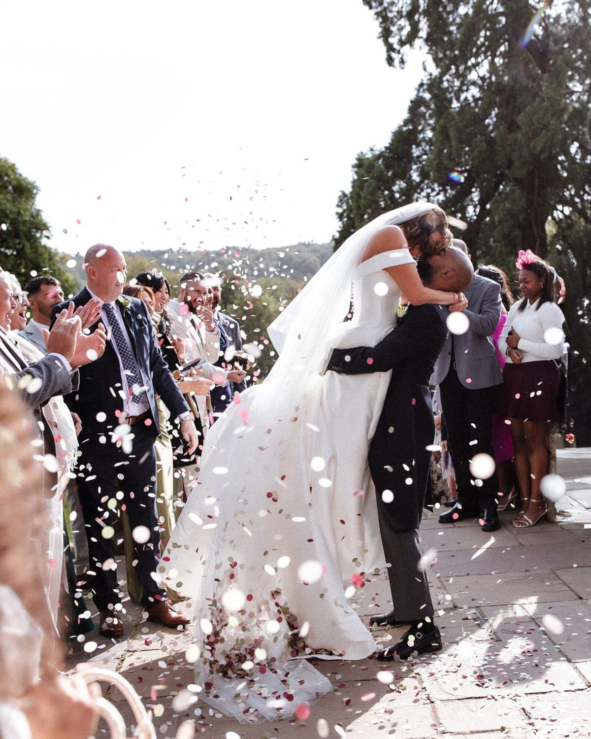 A bride in a white gown kisses and embraces the groom as guests throw flower petals in celebration outdoors, surrounded by trees and sunlight.