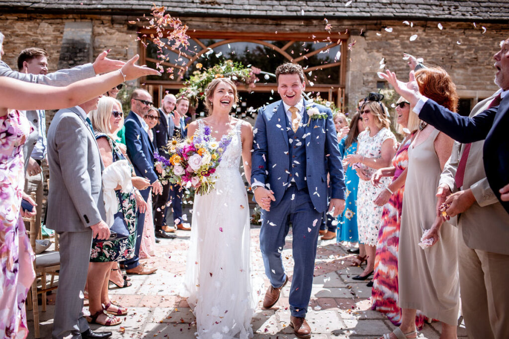 A bride and groom walk hand in hand outside, smiling, as wedding guests toss flower petals in celebration. The bride holds a colorful bouquet, and everyone appears joyful under sunny skies.