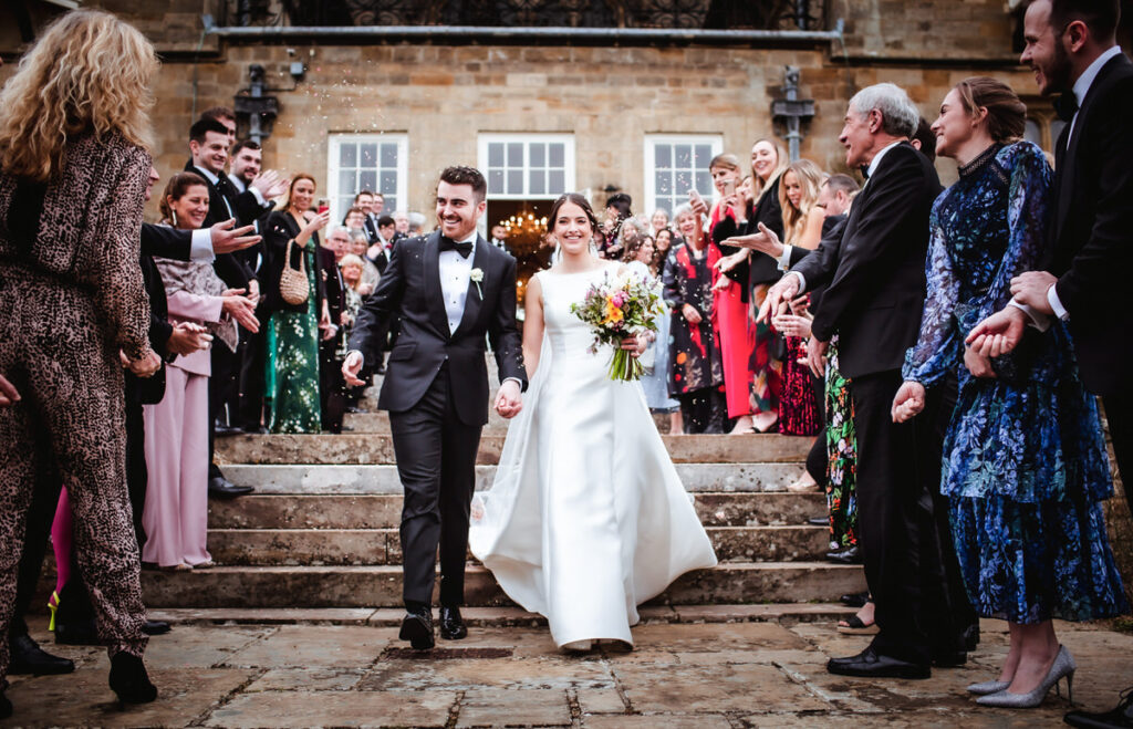 A bride and groom, dressed formally, walk hand in hand down outdoor stone steps, smiling joyfully as guests on both sides cheer and toss confetti. The background shows a large building.