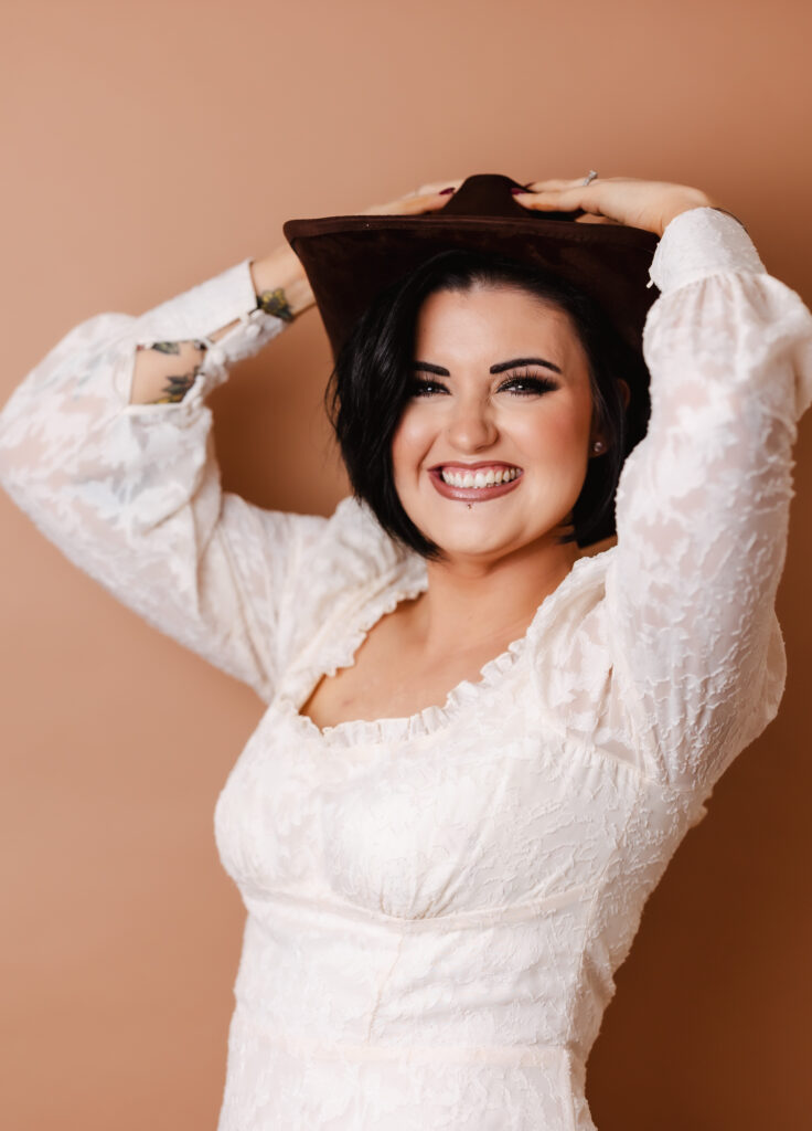 A smiling woman with dark hair in a white textured dress holds a brown hat on her head, standing against a beige background—perfect for Hampshire business headshots with a relaxed, professional vibe.