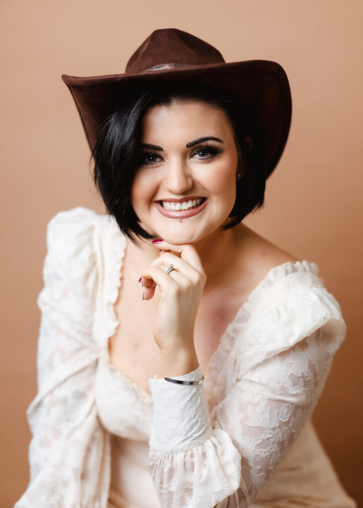 Hampshire headshot photographer. A smiling woman with short dark hair wears a brown cowboy hat and a white lace blouse, posing with her hand under her chin against a beige background—perfect for Hampshire business headshots with a touch of personality.