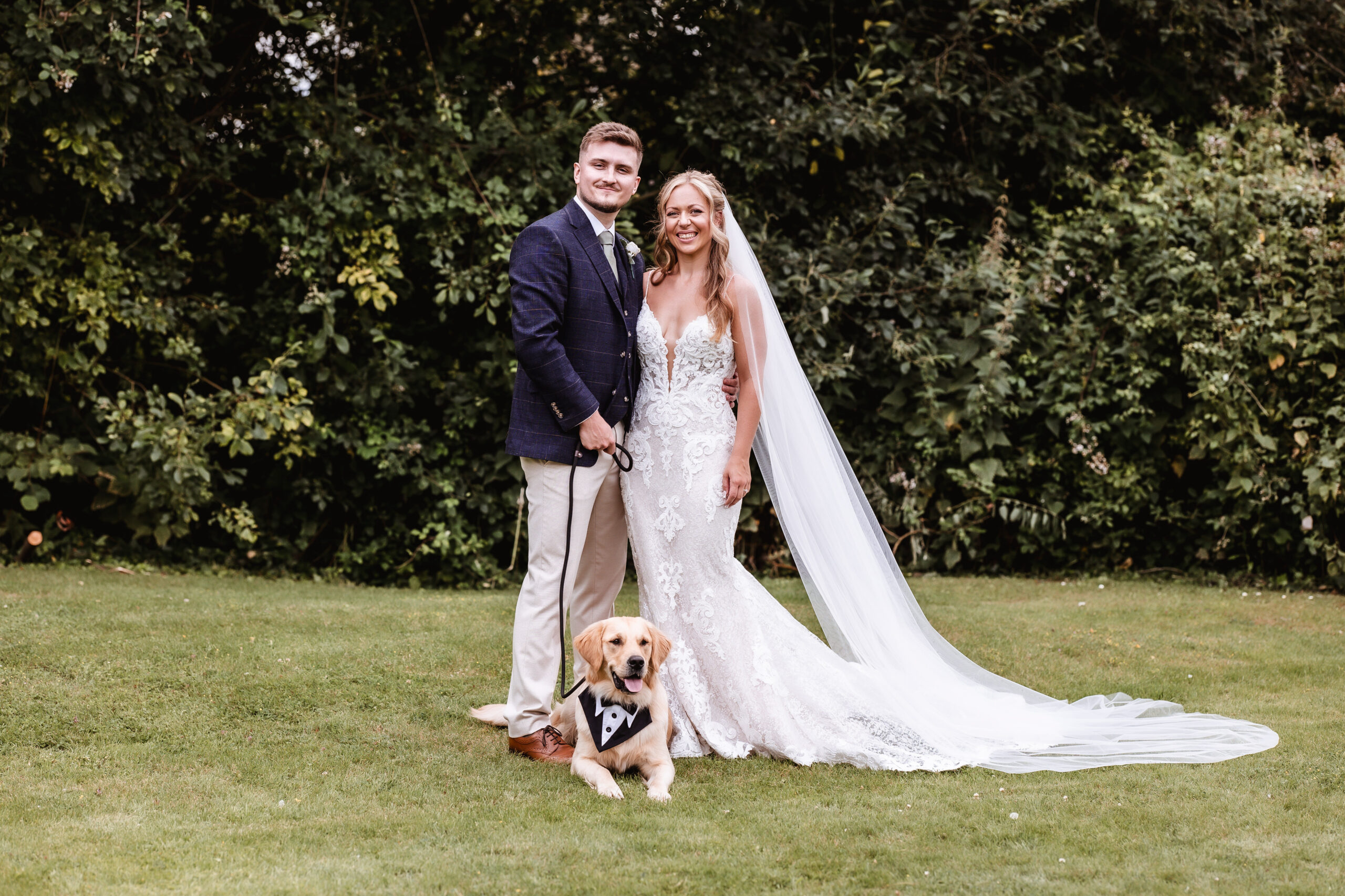 A bride and groom in wedding attire smile on grass before lush trees, with a golden retriever in a bow tie at their feet. The bride’s long, lacy gown and veil complete this charming dog friendly wedding in Hampshire.