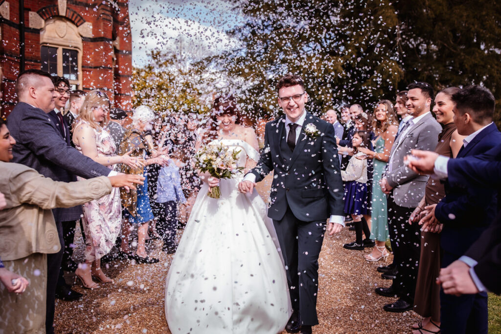 A bride and groom walk outdoors among cheering guests who toss flower petals, celebrating the newlyweds. Everyone is dressed formally, smiling, and standing in front of a red-brick building.