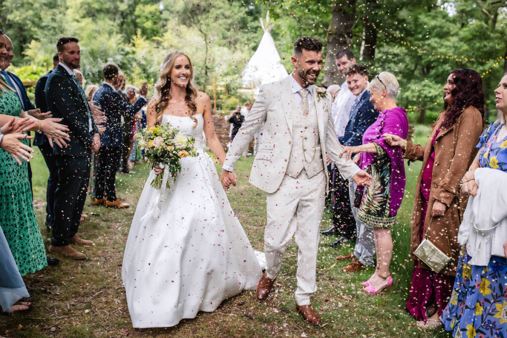 A bride and groom, smiling and holding hands, walk outdoors through guests who are throwing confetti. The scene is festive, with greenery, a white tent in the background, and everyone dressed in formal attire.