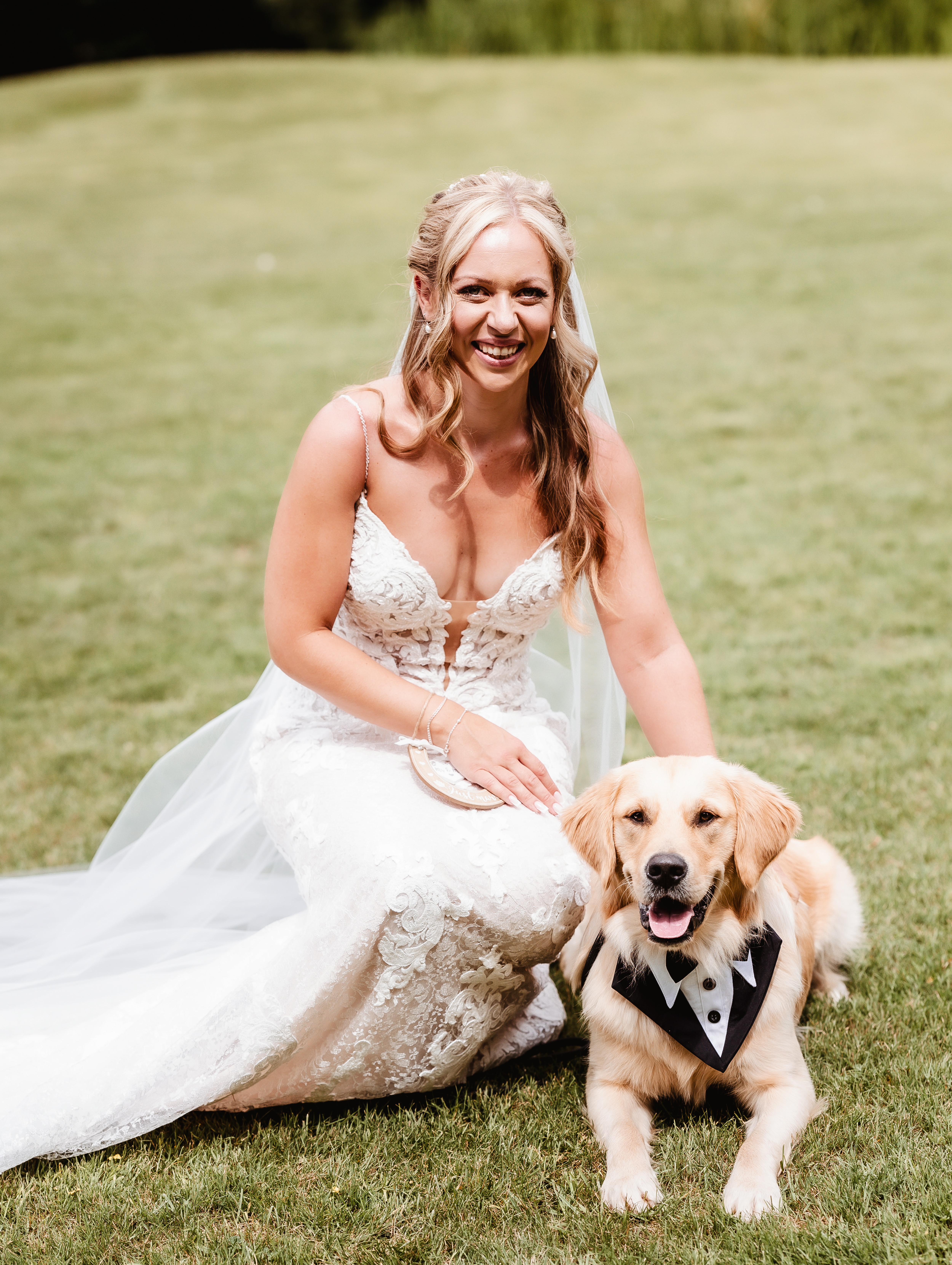 A bride in a white lace wedding dress kneels on grass, smiling and posing with her golden retriever in a tuxedo bandana at a dog friendly wedding in Hampshire.