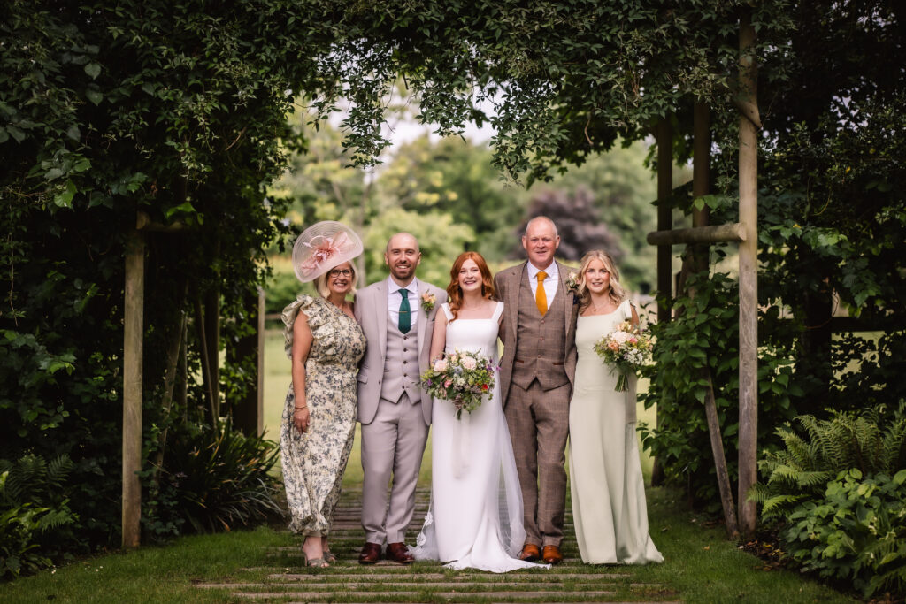 Five people pose outdoors at a wedding, surrounded by greenery. The bride and groom stand in the center, flanked by three others in formal attire. The group is smiling and holding bouquets.