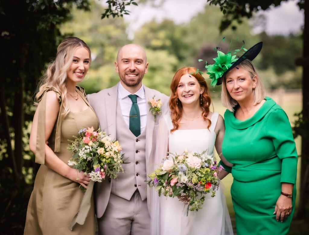 Four people pose outdoors at a wedding: a woman in a gold dress, a man in a beige suit with a green tie, a bride in a white dress, and a woman in a bright green dress with a matching hat. Everyone is smiling and holding flowers.