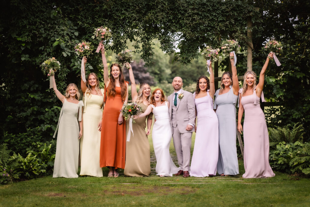 A wedding party stands outdoors on grass, the bride and groom in the center, surrounded by bridesmaids in pastel dresses, all holding bouquets and raising their arms, smiling joyfully under a leafy green arch.