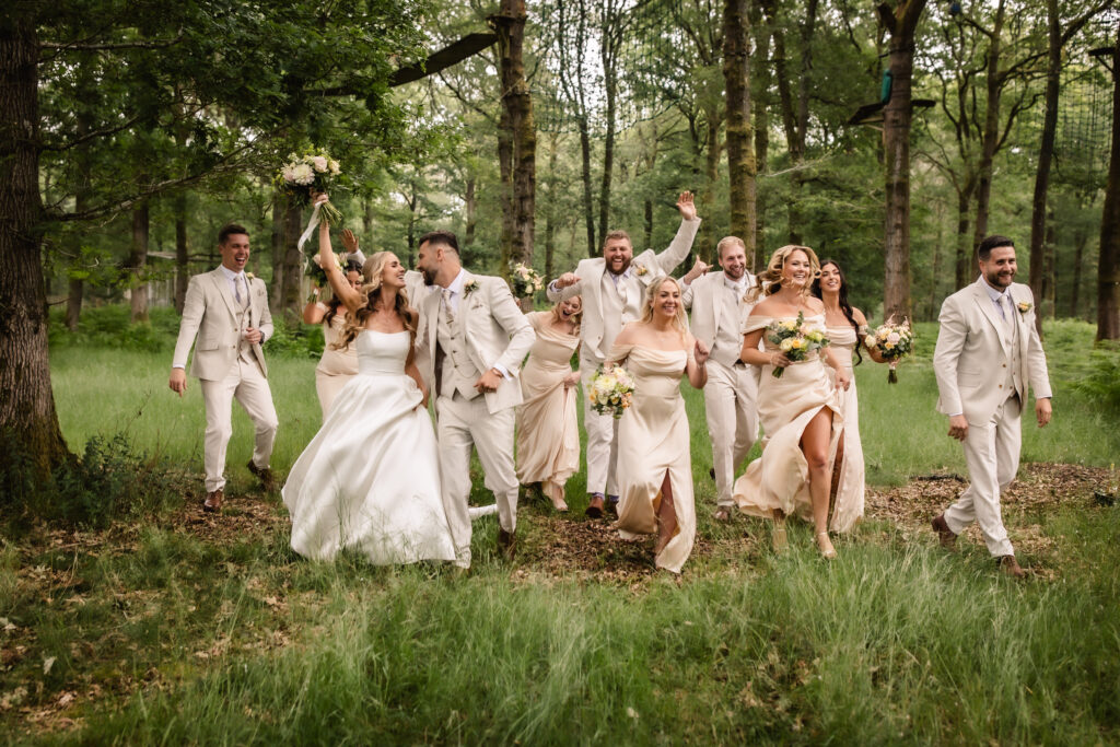 A joyful wedding party, dressed in light-colored formal attire, walks and laughs together through a green, wooded area. The bride and bridesmaids hold bouquets, and the group appears energetic and celebratory.