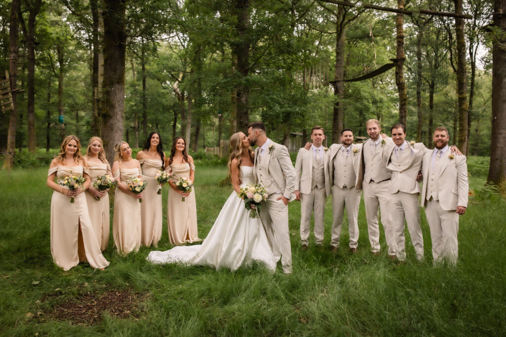 A bride and groom kiss while standing in the center of their wedding party in a lush, green forest. Bridesmaids in light dresses and groomsmen in beige suits smile and pose on either side.