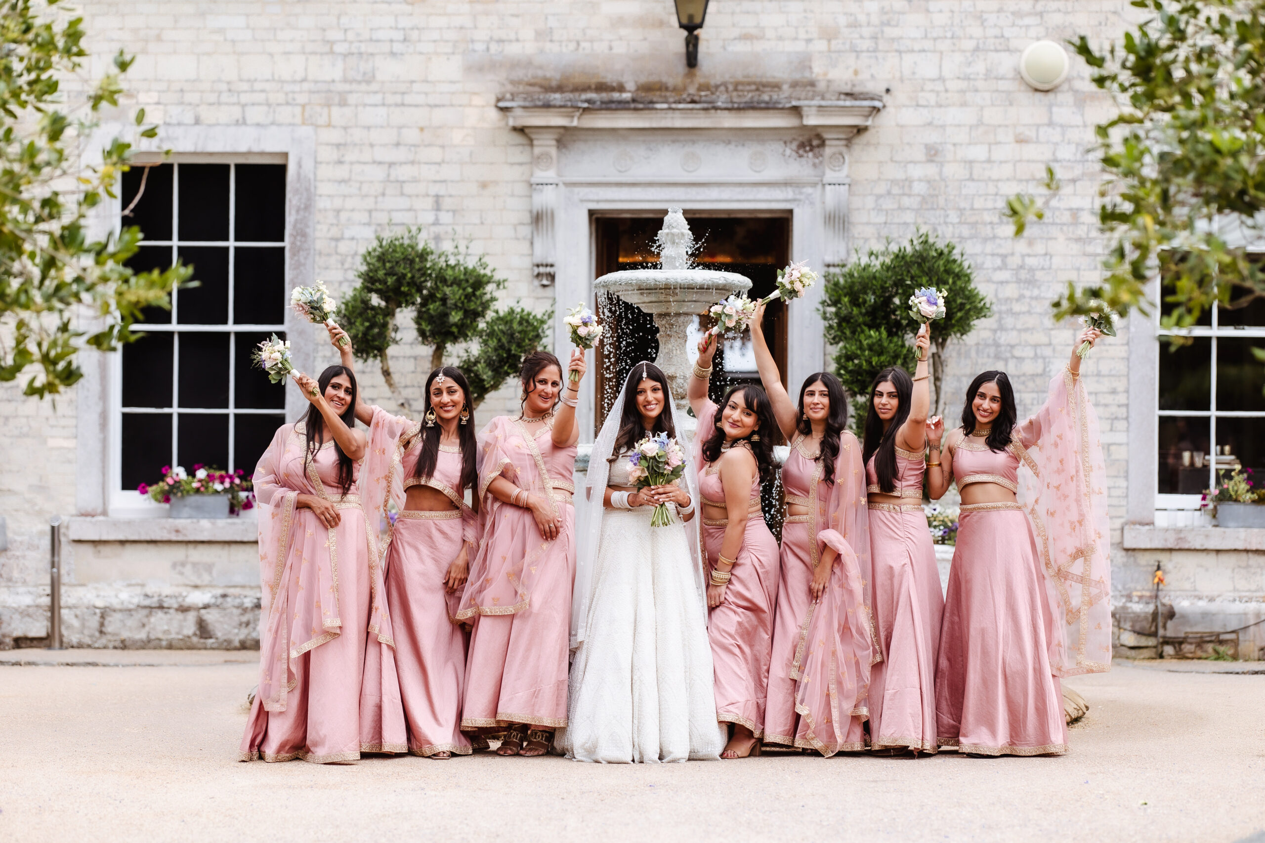 A bride in a white dress and veil stands with eight bridesmaids in matching pink outfits, all smiling and holding bouquets, posing cheerfully in front of a stone building with greenery.
