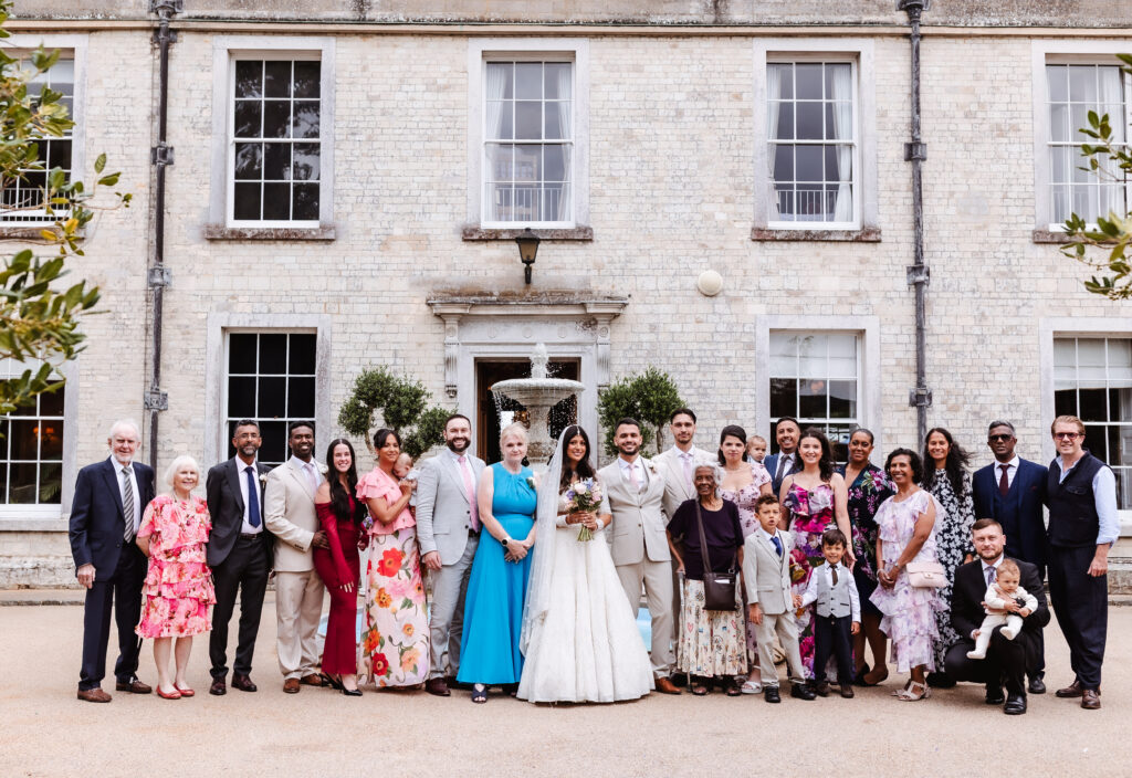 A large wedding group poses in front of a white brick building. The bride and groom stand at the center, surrounded by family and friends dressed in colorful formal attire. Some children and a baby are also present.