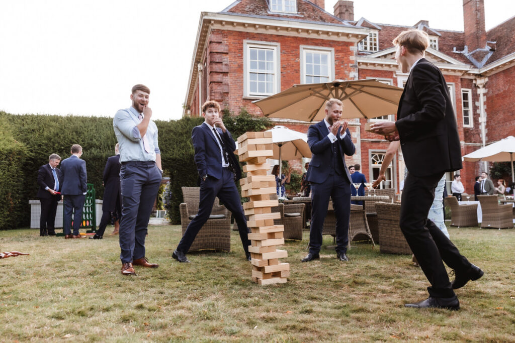 Four men in suits play giant Jenga on a lawn outside a large brick house, with one man catching falling blocks as others react. People and umbrellas are visible in the background.